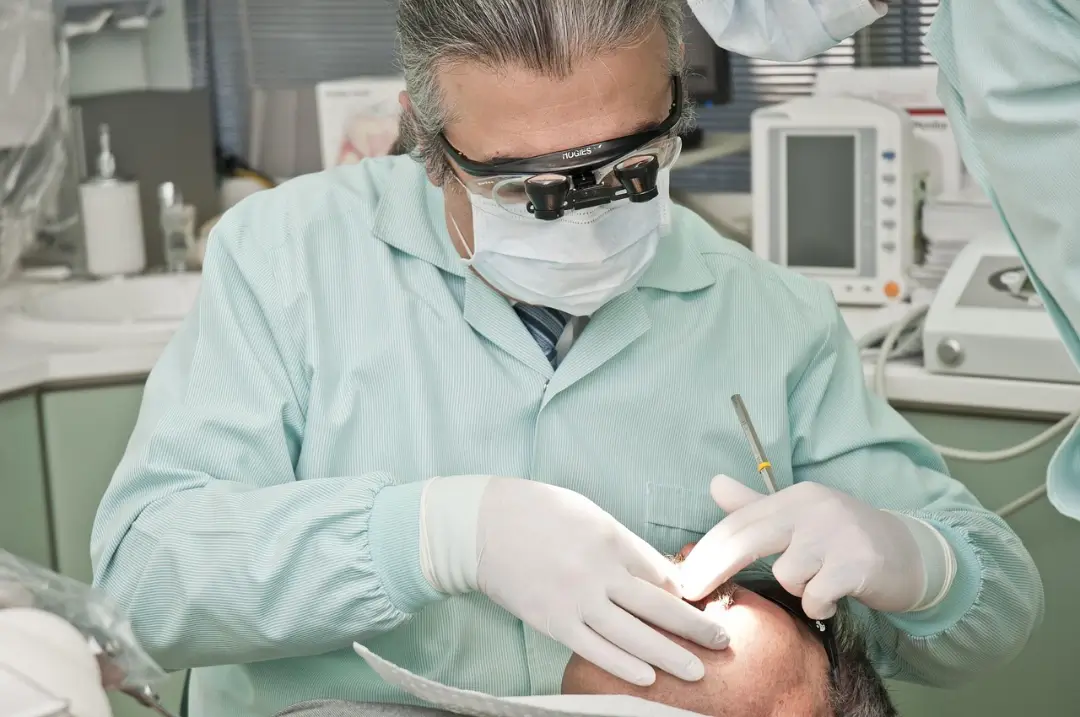 Un dentista con bata verde claro utiliza una herramienta para examinar la cabeza de un paciente en un consultorio dental bien equipado. Una iluminación brillante realza el procedimiento.