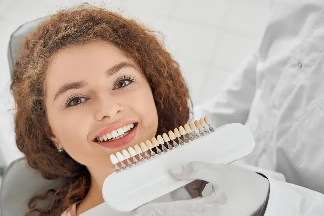 Un profesional dental examina los dientes de un paciente, sosteniendo una paleta de diferentes tonos de dientes para combinar el color.